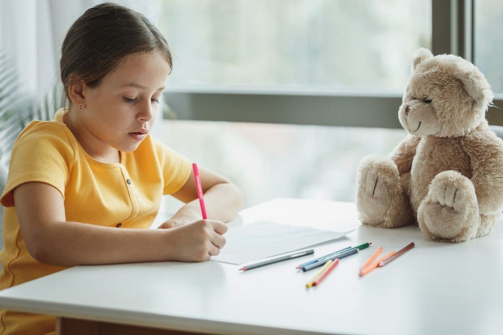 A young girl drawing at a table beside a teddy bear